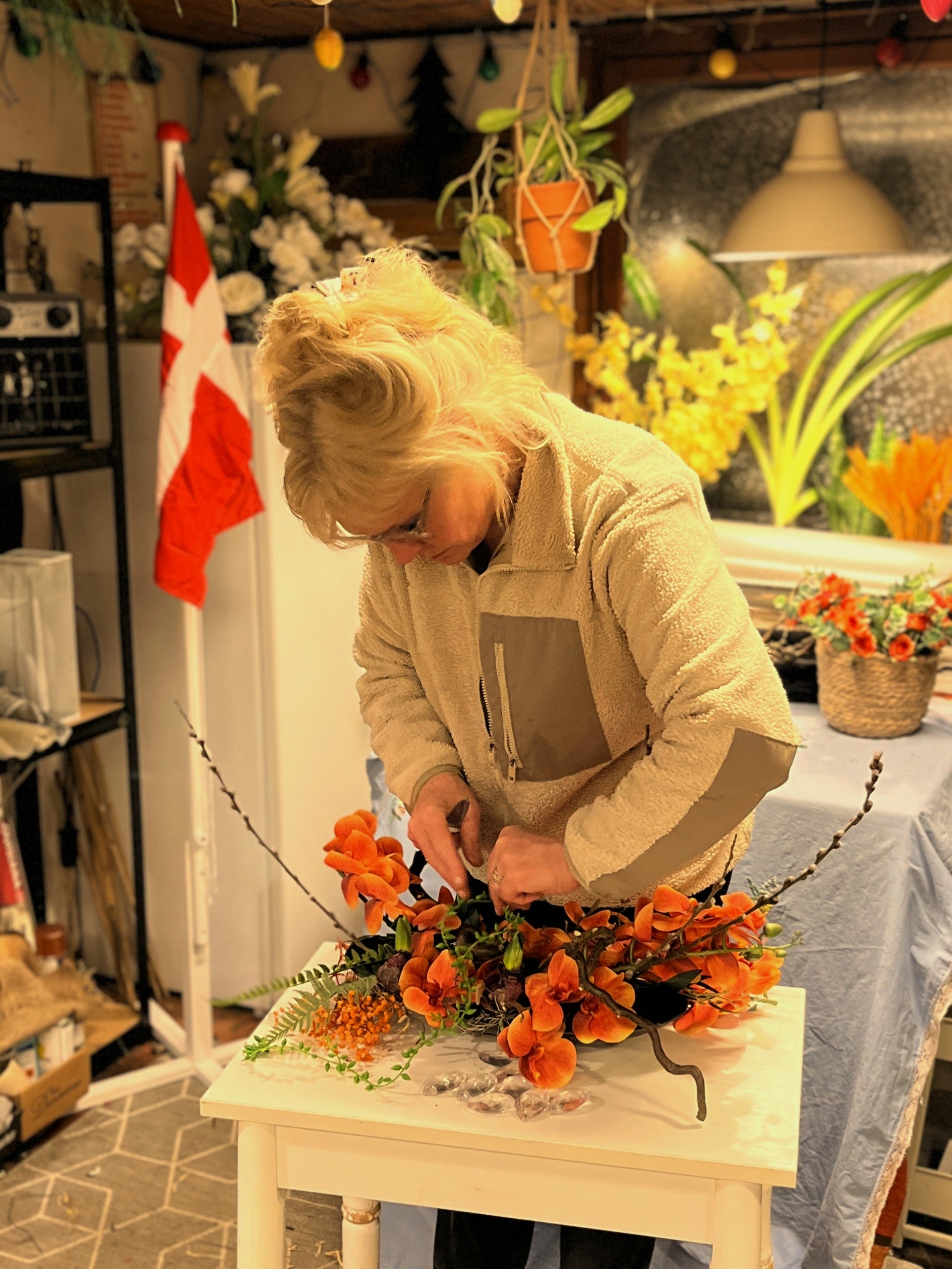 Håndværket bag Linda Blomster. Dekoratør arbejder fokuseret på at færdiggøre en livlig orange orkidédekoration på sit værksted, flankeret af et dansk flag til erhverv.