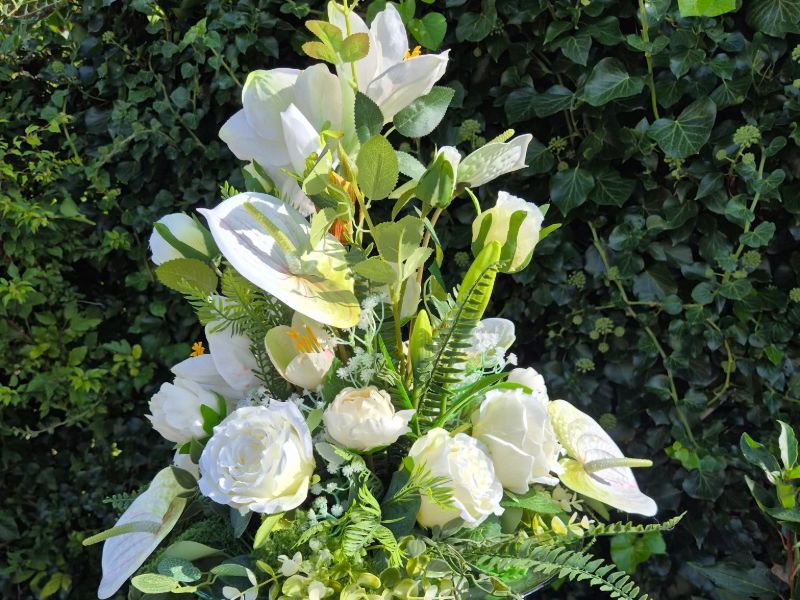 Opstillet hvidt og grønt blomsterarrangement med anthurium, roser og liljer på en lav søjle foran en grøn hæk