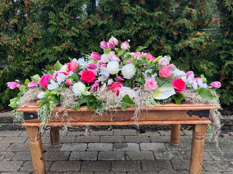 Lavt blomsterarrangement i lyserøde og hvide roser, tulipaner og orkideer med grønt, lagt på et træbord udendørs