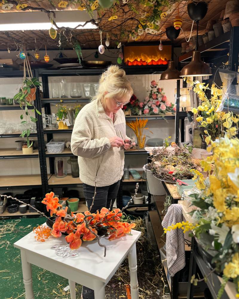 Linda binder blomster ved sit arbejdsbord på værkstedet. Forrest står en dekoration med orange orkideer