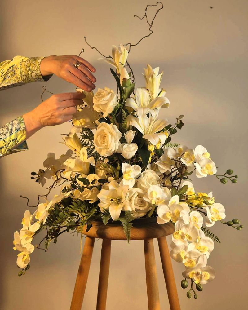 To hænder retter på et stort blomsterarrangement med hvide roser, liljer og orkideer, der står på en træskammel