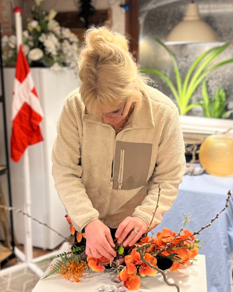 Linda står ved et arbejdsbord og sætter orange orkideer og grene i en dekoration